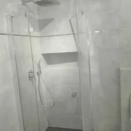 Nowoczesna kabina prysznicowa z deszczownicą i szklanymi drzwiami, wykończona szarą płytką ceramiczną z wnęką na kosmetyki.