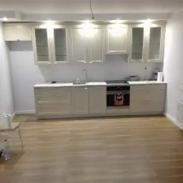 Jasna kuchnia z drewnianą podłogą, białe szafki z przeszklonymi frontami, widoczny fragment grzejnika pod oknem, na pierwszym planie krzesło z rolką papieru i butelką wody.