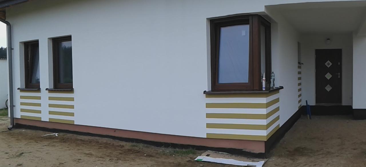 Elewacja domu po malowaniu z poziomymi pasami w kolorze białym i złotym, widoczne okna z brązowymi ramami i butelka silikonu na parapecie.