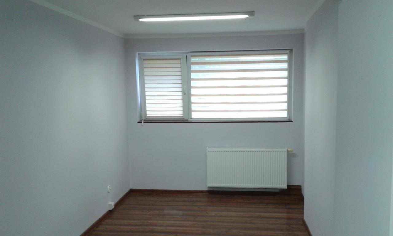 Pomalowane na szaro wnętrze pokoju z drewnianą podłogą, białym grzejnikiem i oknem z żaluzjami w biało-brązowe pasy.