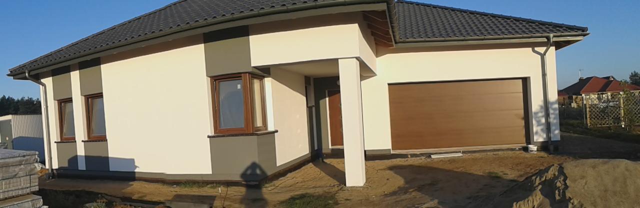 Nowoczesny dom jednorodzinny z kremową elewacją, szarymi akcentami i brązową bramą garażową, widok z zewnątrz na tle błękitnego nieba i zieleni drzew.