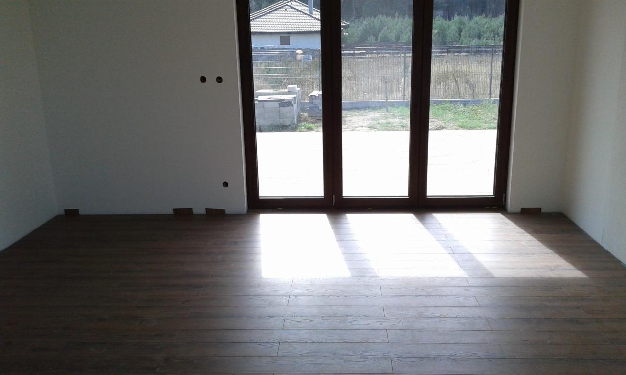 Nowo ułożona, ciemna podłoga panelowa w pokoju z dużym, trzyskrzydłowym oknem balkonowym; widoczne przygotowane do montażu listwy przypodłogowe.