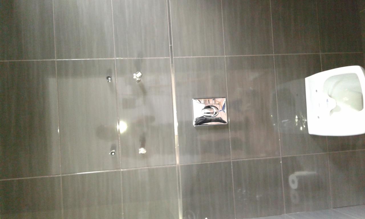 Szare płytki ścienne z zamontowaną białą toaletą i chromowanym przyciskiem spłukiwania; widoczne otwory po wierceniu.