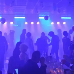 Impreza taneczna z oświetleniem scenicznym, lustrzanymi kulami i sylwetkami ludzi w tle oraz osobą nalewającą napój w pierwszym planie.