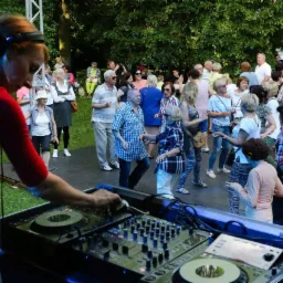 Kobieta DJ z niebieskimi włosami miksuje muzykę na konsoli DJ-ską na plenerowej imprezie tanecznej, w tle bawiący się tłum ludzi i drzewa.