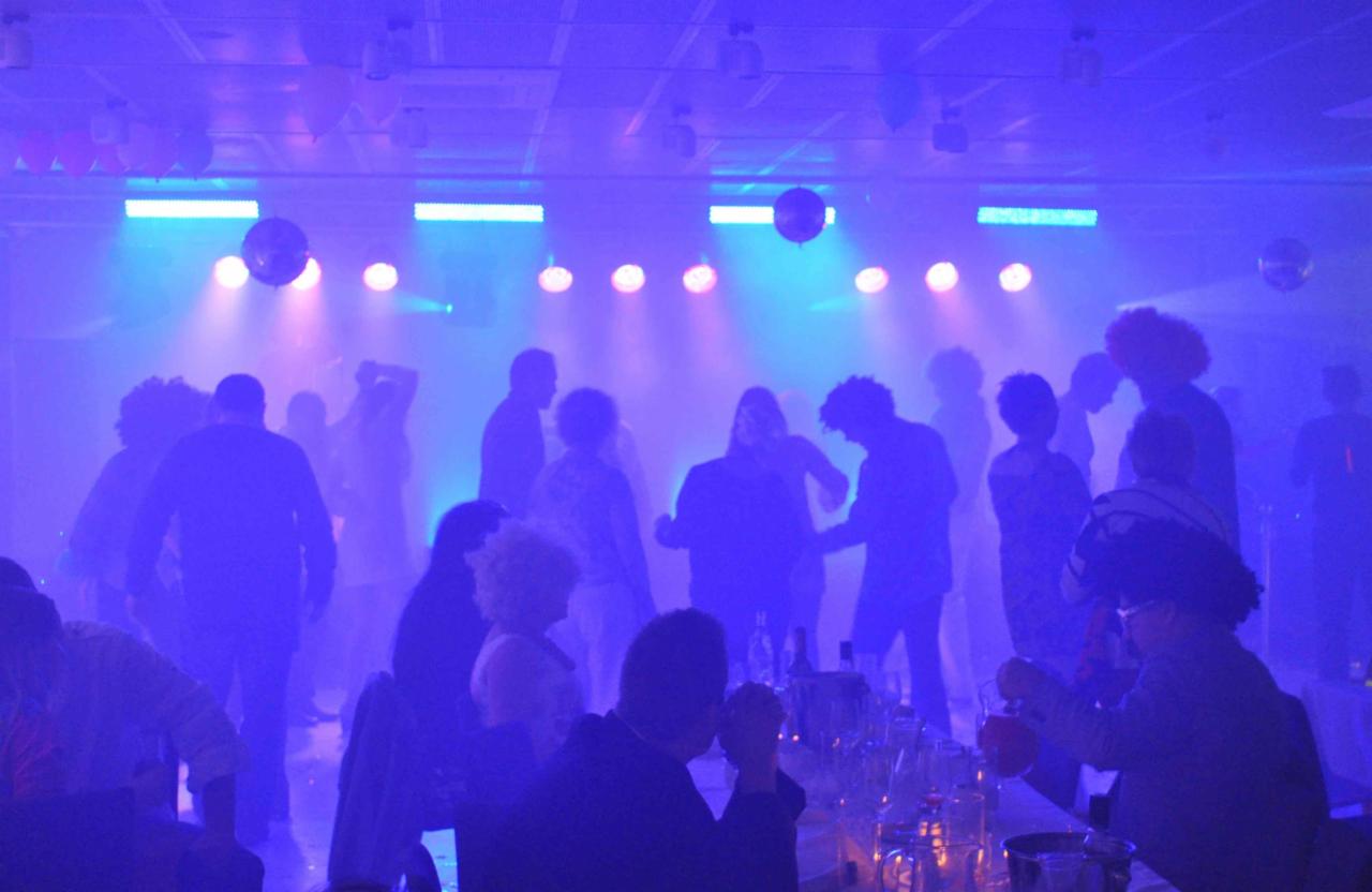 Impreza taneczna z oświetleniem scenicznym, lustrzanymi kulami i sylwetkami ludzi w tle oraz osobą nalewającą napój w pierwszym planie.