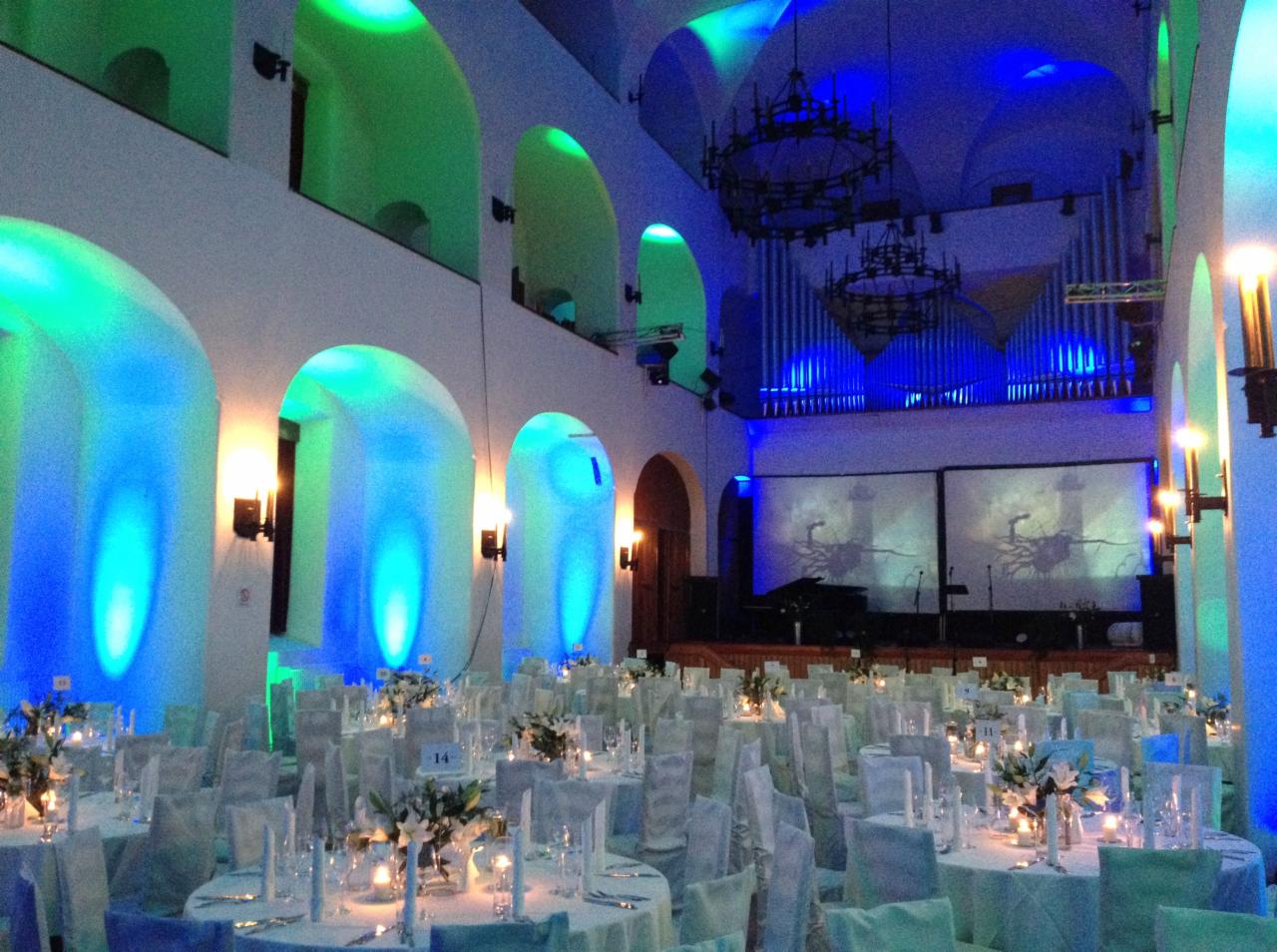 Uroczysta sala balowa z okrągłymi stołami nakrytymi białymi obrusami, ozdobionymi kwiatami i świecami, oświetlona zielonymi i niebieskimi światłami, z organami i ekranami projekcyjnymi w tle.