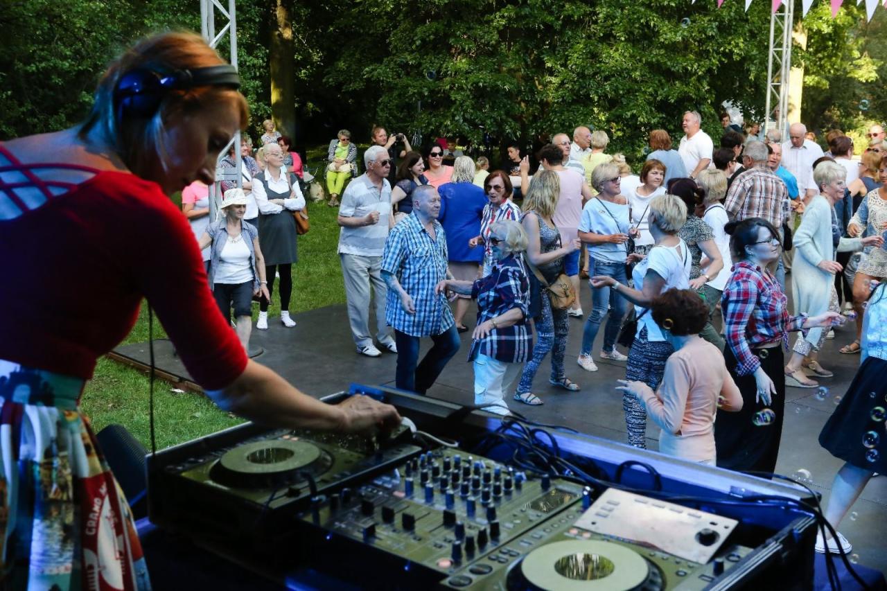 Kobieta DJ z niebieskimi włosami miksuje muzykę na konsoli DJ-ską na plenerowej imprezie tanecznej, w tle bawiący się tłum ludzi i drzewa.