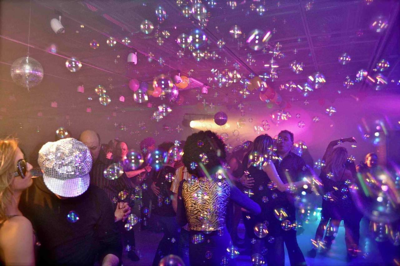 Roztańczony tłum w dyskotekowym oświetleniu, wypełniony unoszącymi się bańkami mydlanymi, z lustrzanymi kulami i balonami na suficie.