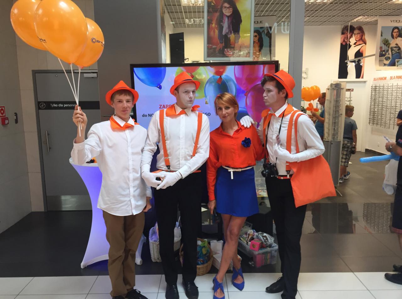 Grupa osób w pomarańczowych kapeluszach i z białym makijażem pozuje z pomarańczowymi balonami w sklepie, prawdopodobnie podczas wydarzenia promocyjnego.