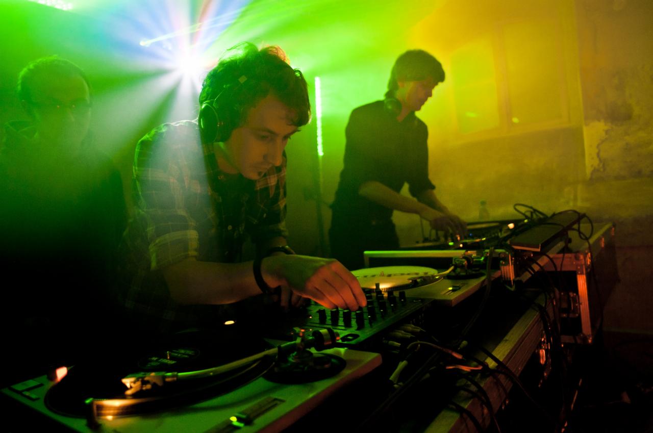 Dwóch DJ-ów z słuchawkami gra muzykę na gramofonach podczas imprezy w Szczecinie, oświetlonych zielonym i żółtym światłem.