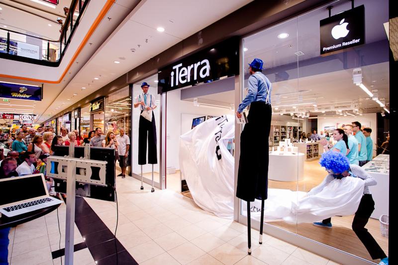 Otwarcie sklepu iTerra w centrum handlowym; mężczyźni na szczudłach w niebieskich kapeluszach i szelkach, rozwijają białe płótno z logo, tłum widzów.