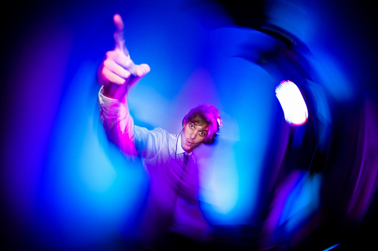 Rozmazane ujęcie DJ-a z podniesionym palcem, w słuchawkach, na tle intensywnego niebiesko-fioletowego oświetlenia scenicznego.