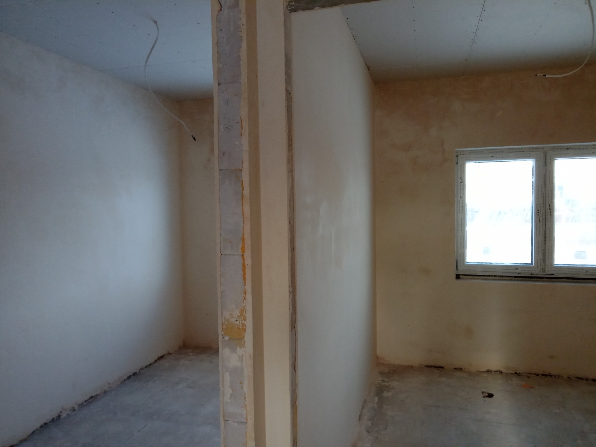 Wnętrze w trakcie prac wykończeniowych: ściany po nałożeniu gładzi, widoczne fragmenty sufitu z płyt kartonowo-gipsowych oraz surowy beton na podłodze.