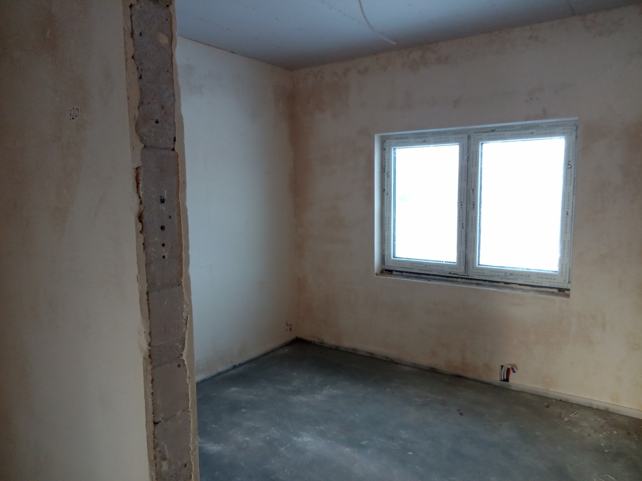 Surowy stan wnętrza z oknem: ściany po tynkowaniu, widoczne niedoskonałości i przygotowanie do dalszych prac wykończeniowych, surowa wylewka na podłodze, odsłonięte przewody instalacji w ścianie.