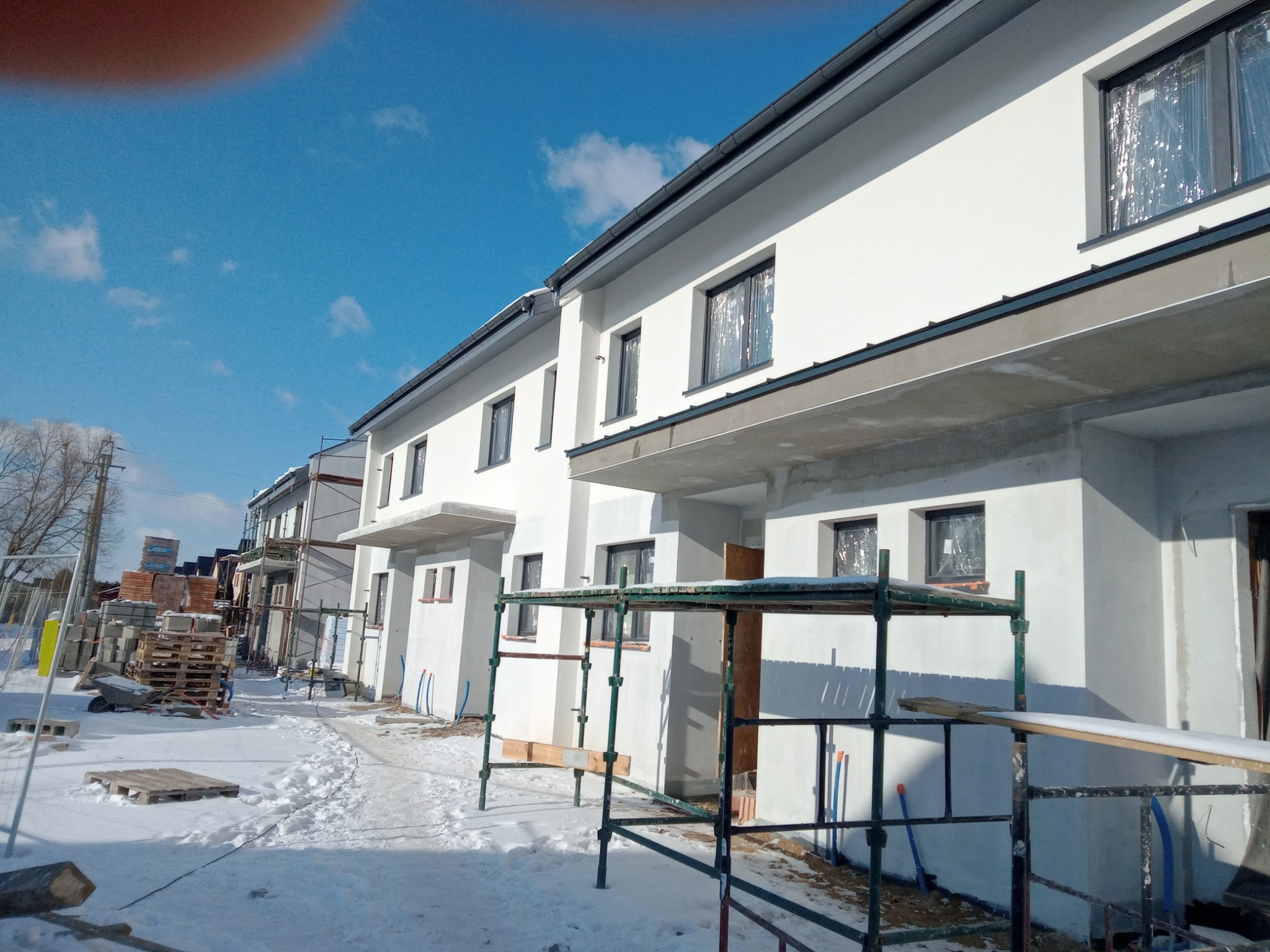 Nowe, otynkowane budynki w zabudowie szeregowej zimą, z widocznymi rusztowaniami i materiałami budowlanymi na paletach.