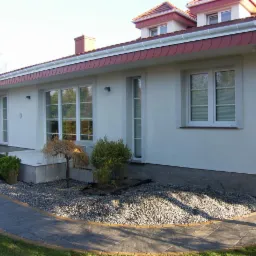 Jasny dom jednorodzinny z wykończoną elewacją, widoczne okna z białymi ramami, czerwony dach i ozdobne krzewy przed budynkiem.