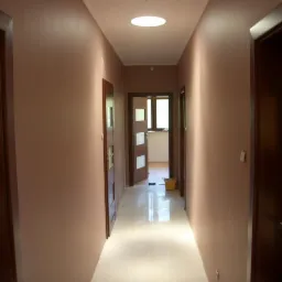 Wnętrze korytarza po malowaniu ścian na kolor brudnego różu, widoczne ciemne drewniane drzwi z przeszkleniami i okrągłe okno w suficie, podłoga wyłożona jasnymi płytkami, w tle żółte wiadro.