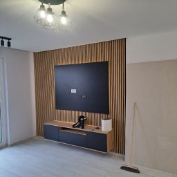 KAROL KOŁSUT - Nowoczesny salon z drewnianą ścianą z lameli, podwieszaną szafką RTV i czarnym panelem TV. Na podłodze jasne panele. Widoczna lampa sufitowa i częściowo ściana do wykończenia.