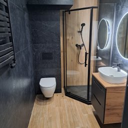 KAROL KOŁSUT - Nowoczesna łazienka z kabiną prysznicową, umywalką na szafce i toaletą. Ciemne płytki imitujące marmur kontrastują z drewnopodobną podłogą i ścianą w kabinie.