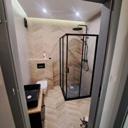 KAROL KOŁSUT - Nowoczesna łazienka po remoncie: kabina prysznicowa typu walk-in z czarnymi ramami, umywalka nablatowa i płytki imitujące drewno ułożone w jodełkę.