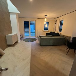 KAROL KOŁSUT - Salon po wykończeniu: parkiet w jodełkę, nowoczesne meble, lampy wiszące. Widok na okno z żaluzjami i wyjście na zewnątrz. Ciepłe oświetlenie.