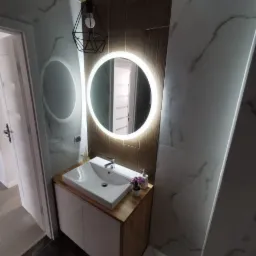 Nowoczesna łazienka z okrągłym lustrem podświetlanym LED, białą umywalką nablatową na szafce z drewnianym blatem, marmurowe płytki na ścianach i wisząca lampa z czarnym, geometrycznym kloszem.