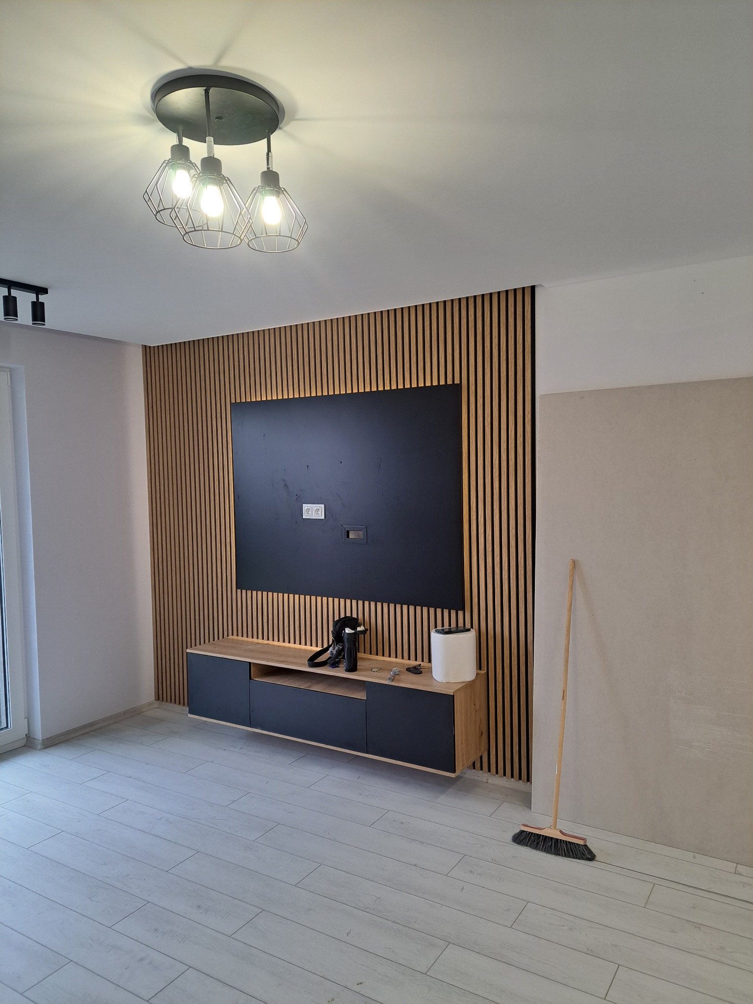 Nowoczesny salon z drewnianą ścianą z lameli, podwieszaną szafką RTV i czarnym panelem TV. Na podłodze jasne panele. Widoczna lampa sufitowa i częściowo ściana do wykończenia.