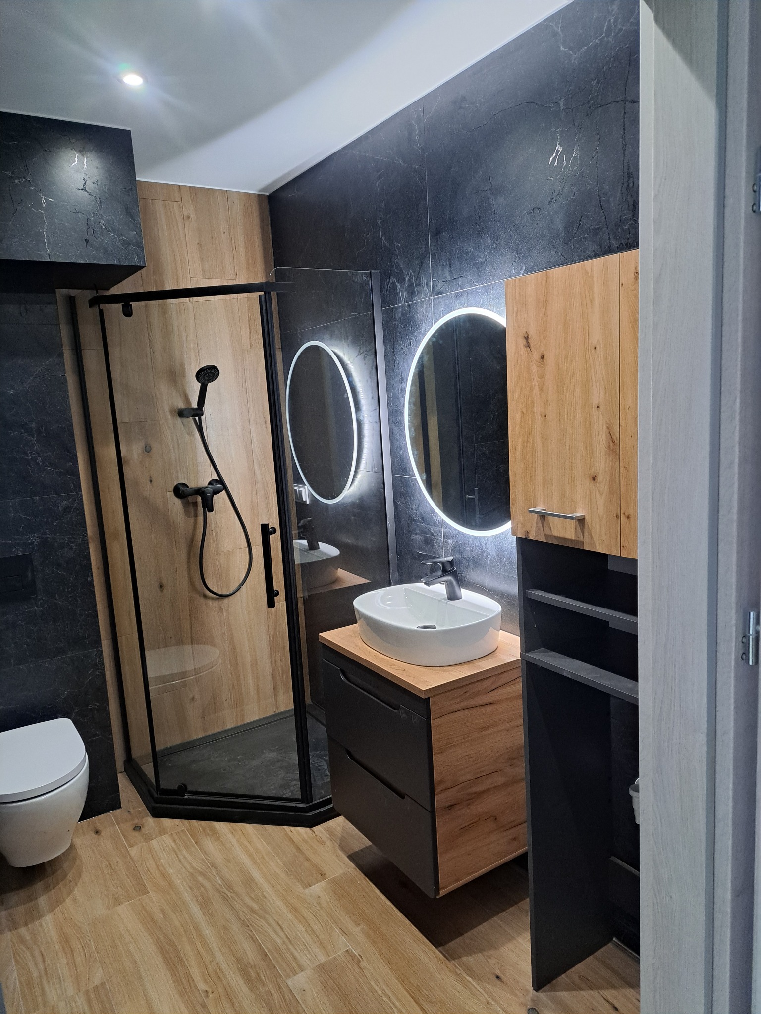Nowoczesna łazienka z kabiną prysznicową, umywalką z szafką i okrągłym lustrem z podświetleniem LED. Czarno-drewniane wykończenie, jasna podłoga.