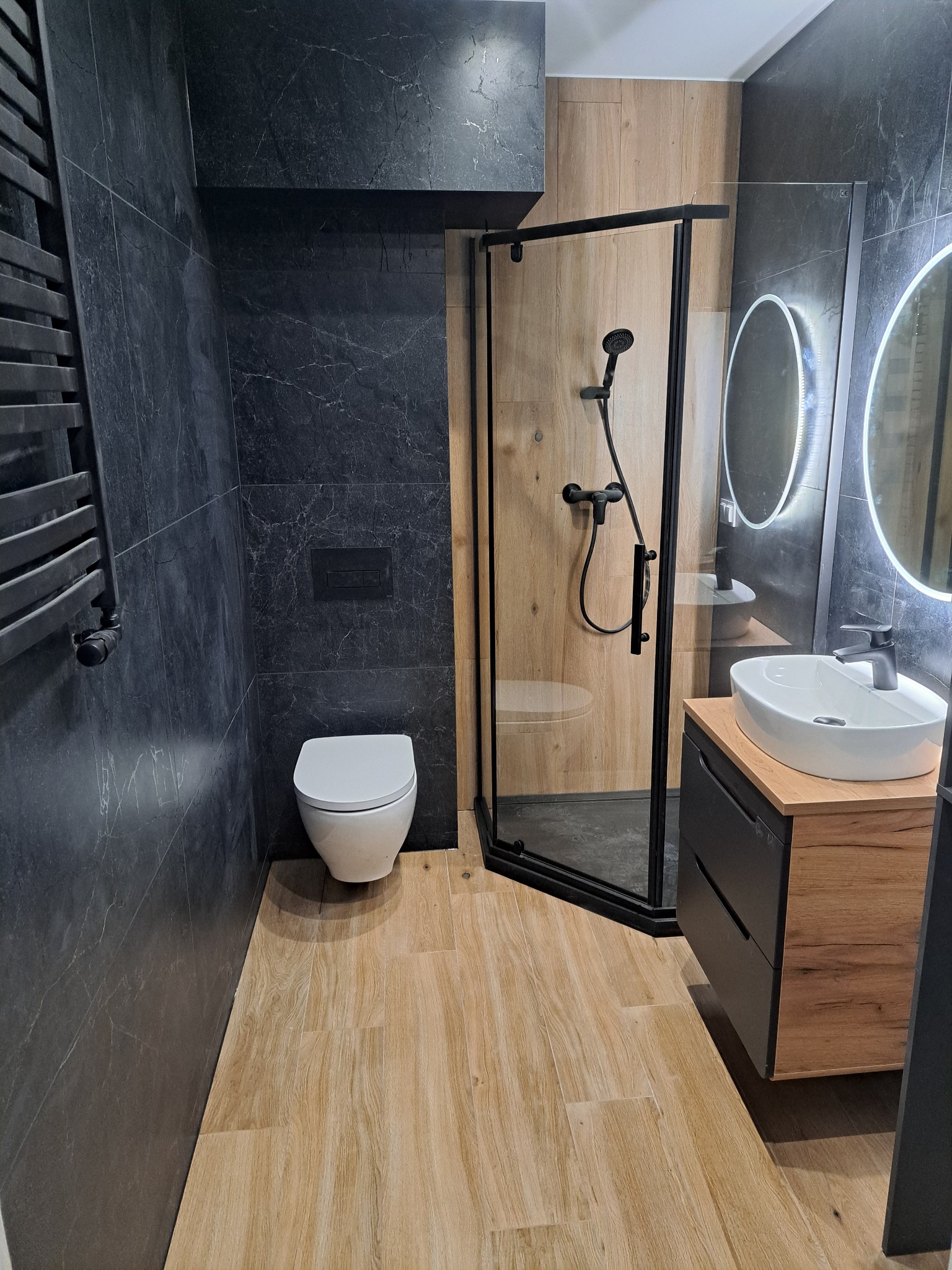 Nowoczesna łazienka z kabiną prysznicową, umywalką na szafce i toaletą. Ciemne płytki imitujące marmur kontrastują z drewnopodobną podłogą i ścianą w kabinie.
