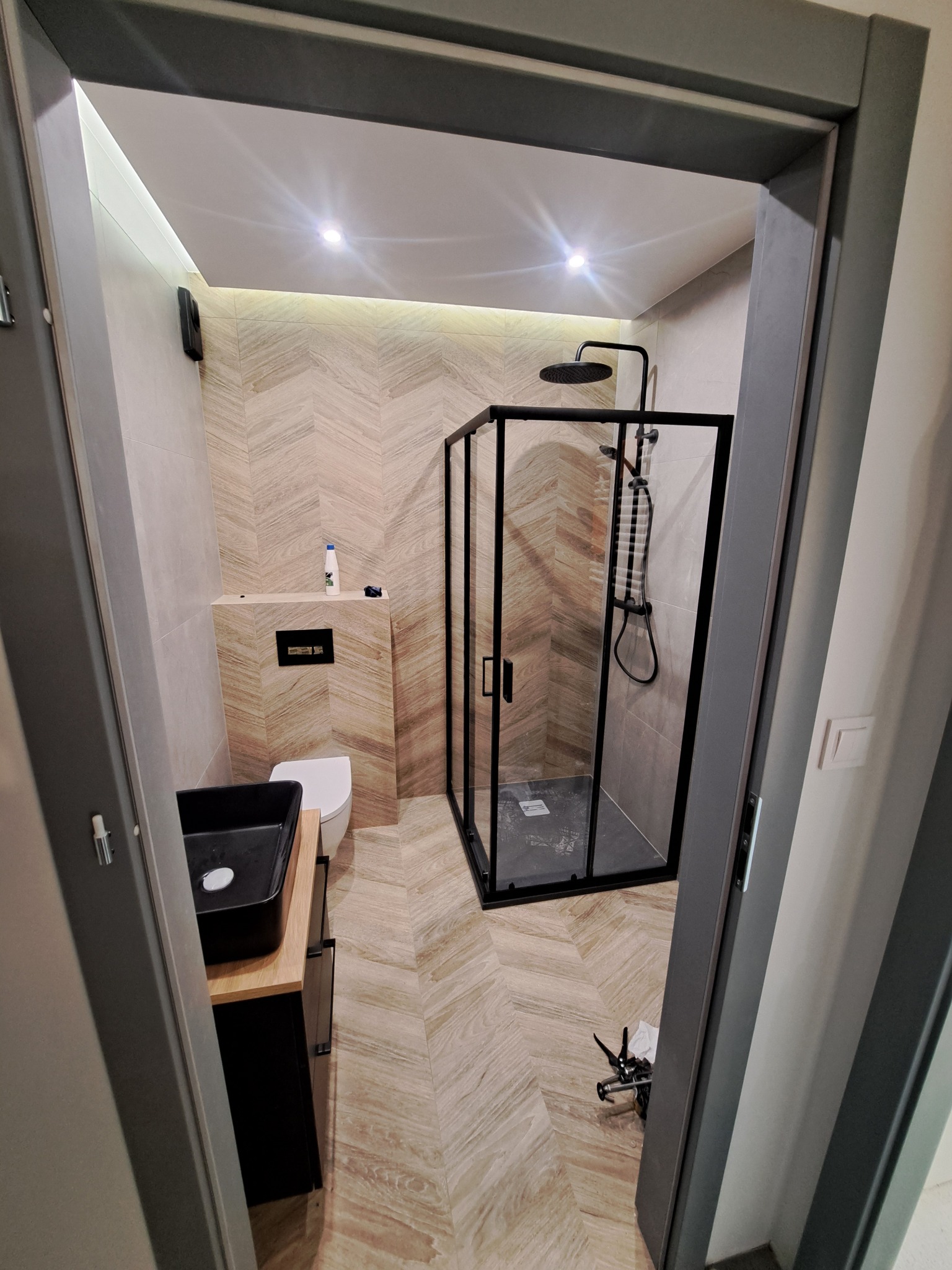 Nowoczesna łazienka po remoncie: kabina prysznicowa typu walk-in z czarnymi ramami, umywalka nablatowa i płytki imitujące drewno ułożone w jodełkę.