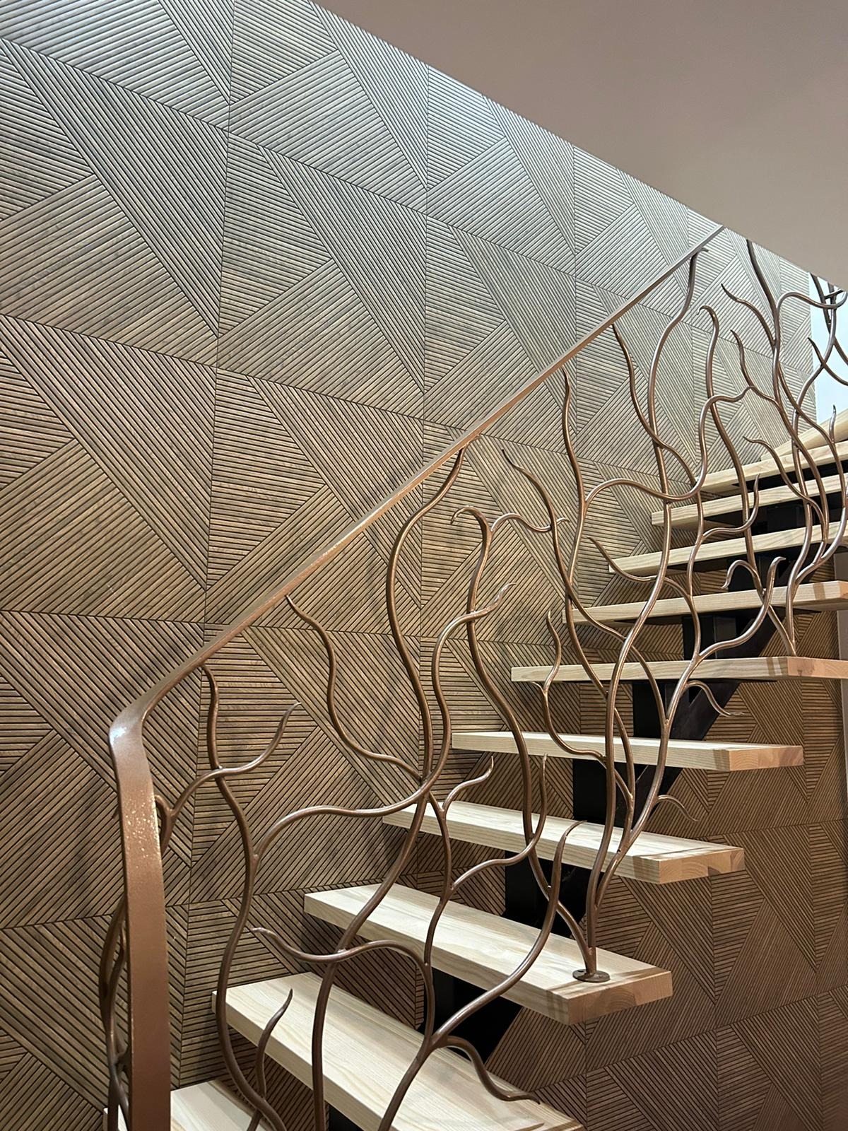 Schody z drewnianymi stopniami i metalową balustradą o organicznym wzorze na tle ściany z dekoracyjnymi panelami w geometryczny wzór.