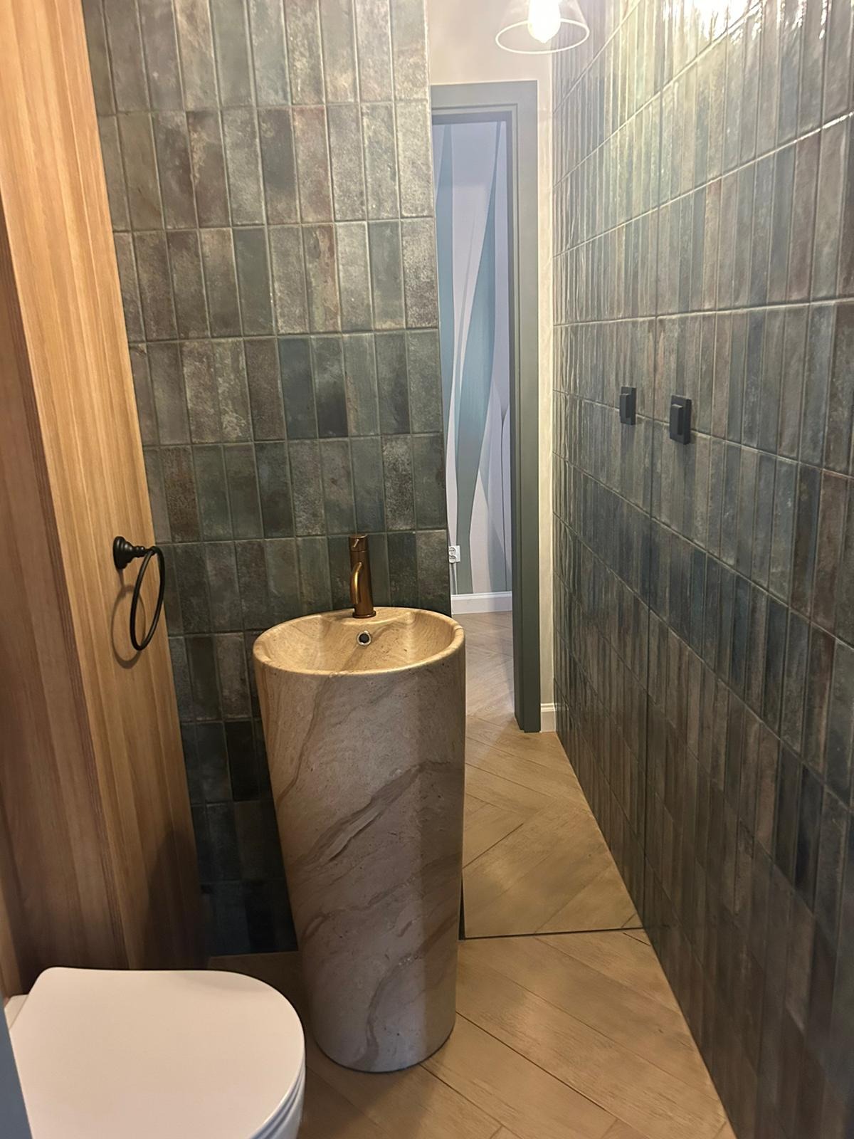 Stylowa łazienka z umywalką w kształcie kolumny, ciemnymi płytkami na ścianach i drewnianymi elementami wykończenia. Widoczny fragment toalety i drzwi w tle.