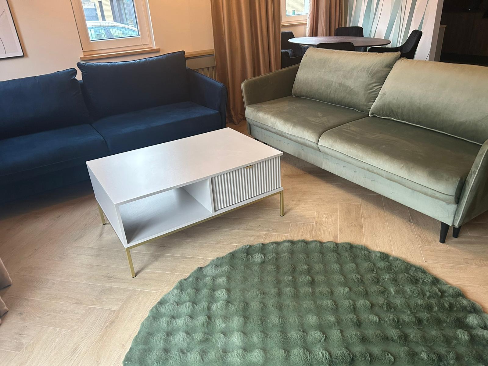 Stylowy salon z dwoma sofami: granatową i oliwkową. Biały stolik kawowy z ryflowanym frontem i okrągły, zielony dywan. Parkiet w jodełkę i beżowe zasłony w tle.