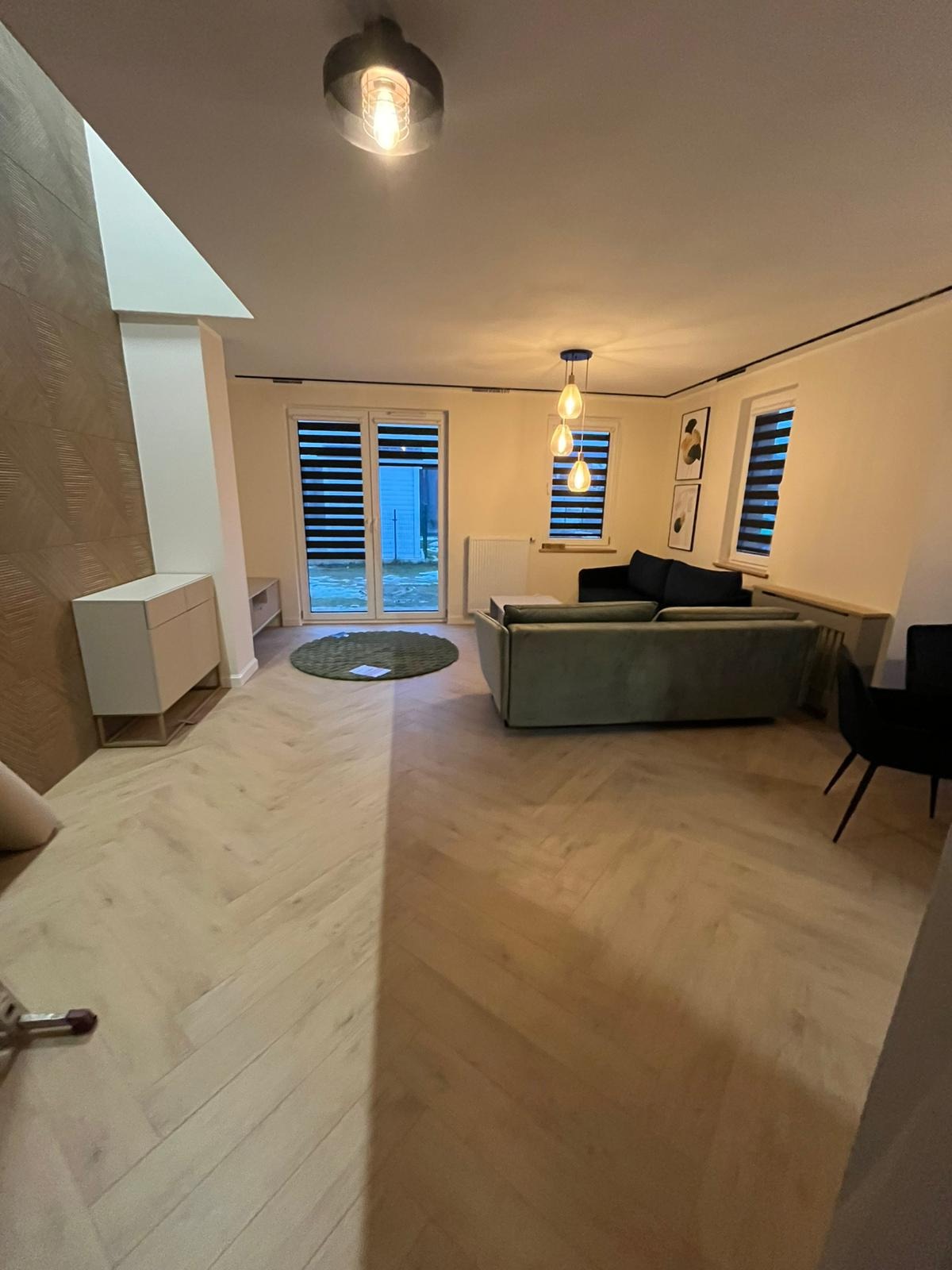 Salon po wykończeniu: parkiet w jodełkę, nowoczesne meble, lampy wiszące. Widok na okno z żaluzjami i wyjście na zewnątrz. Ciepłe oświetlenie.