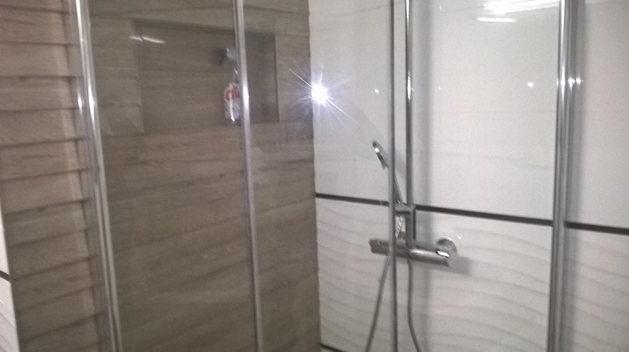 Szklana kabina prysznicowa z chromowaną armaturą na tle ściany wyłożonej białymi i szarymi płytkami z delikatnym wzorem fal.