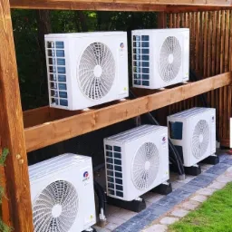 Cztery zewnętrzne jednostki klimatyzacyjne marki Gree zamontowane na drewnianej konstrukcji w ogrodzie, widoczne przewody i logo firmy Smart Dom Klima.