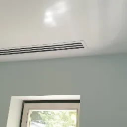 Nowoczesny, liniowy nawiewnik sufitowy z czarnymi kratkami w jasnym pomieszczeniu, widoczny również mały, prostokątny otwór wentylacyjny oraz fragment okna.