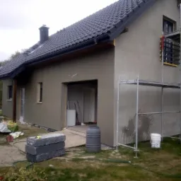 Dom jednorodzinny w trakcie prac elewacyjnych, widoczne rusztowanie, świeżo nałożona warstwa tynku oraz materiały budowlane na trawniku.