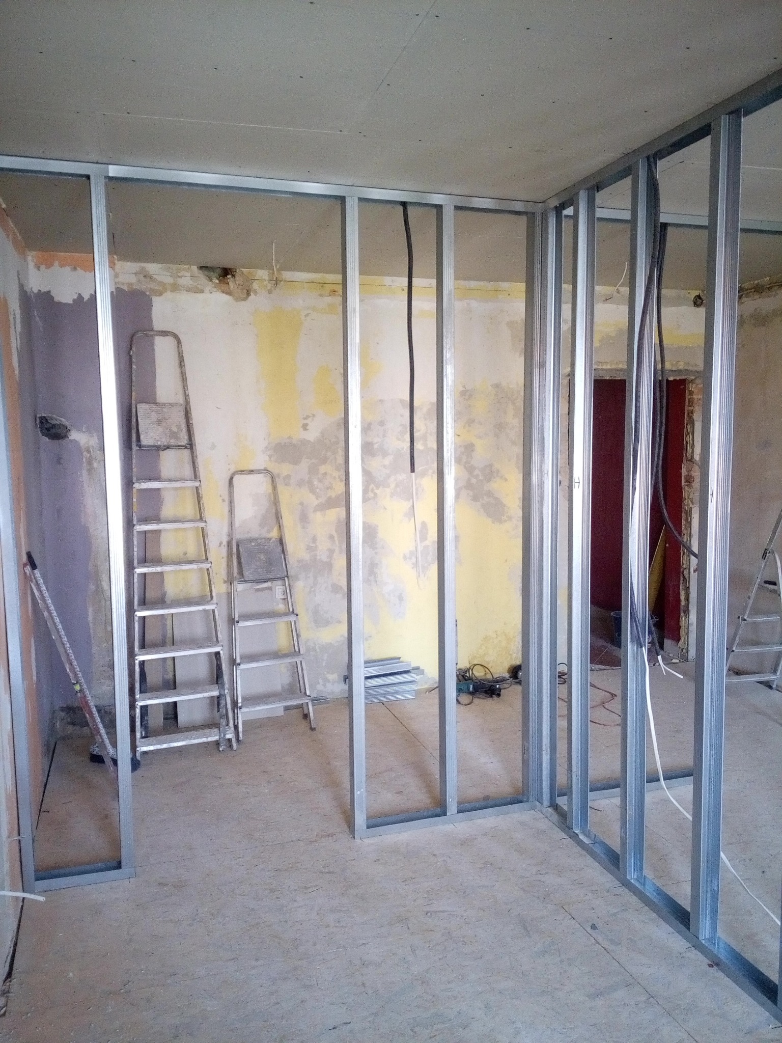 Konstrukcja szkieletowa z profili metalowych do ścian działowych w trakcie montażu, widoczne dwie drabiny aluminiowe, przewody elektryczne oraz fragmenty starego tynku na ścianach.