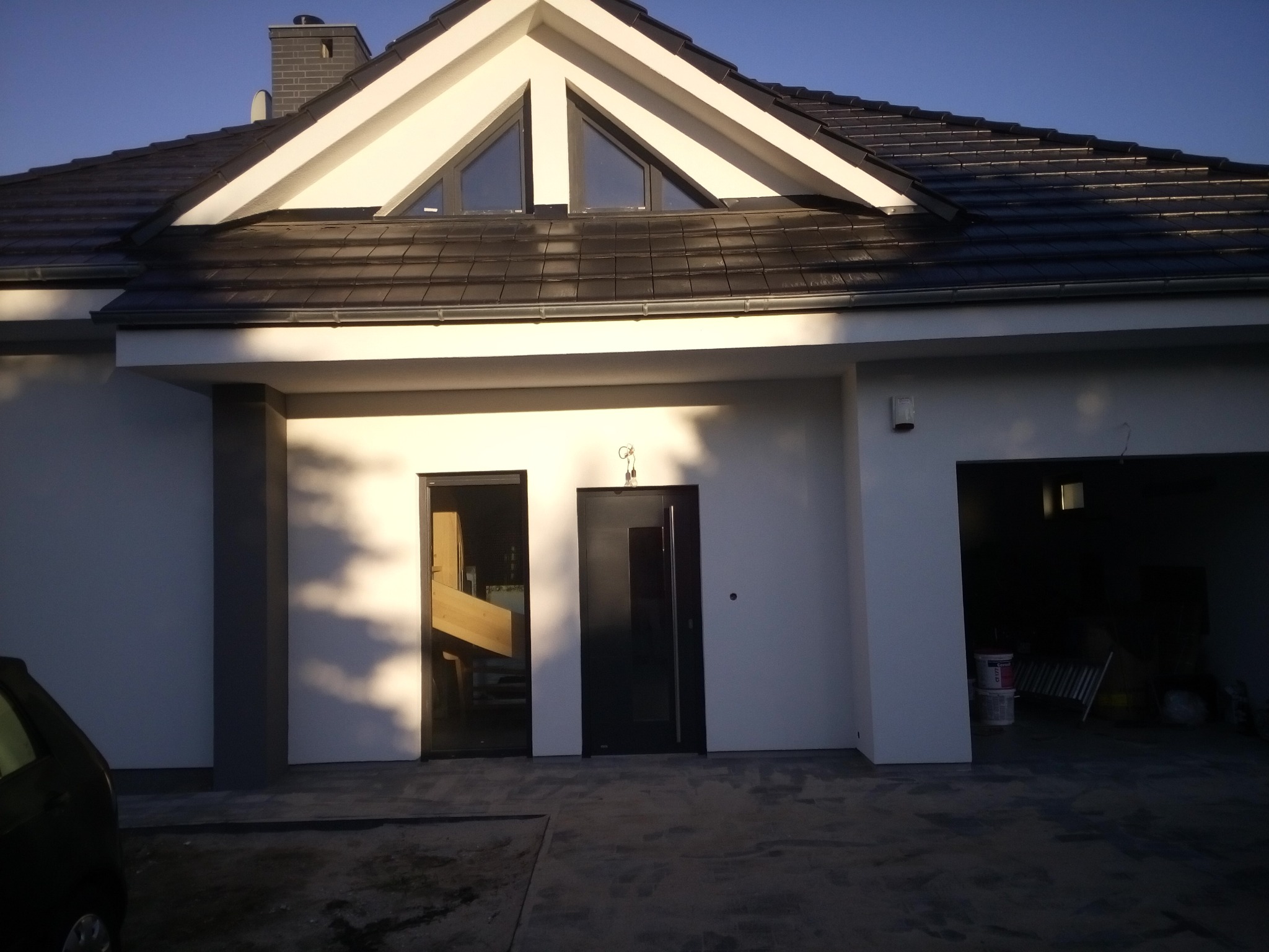 Nowoczesny dom jednorodzinny z białymi ścianami, ciemną dachówką i antracytowymi oknami oraz drzwiami wejściowymi, widziany z zewnątrz w słoneczny dzień.