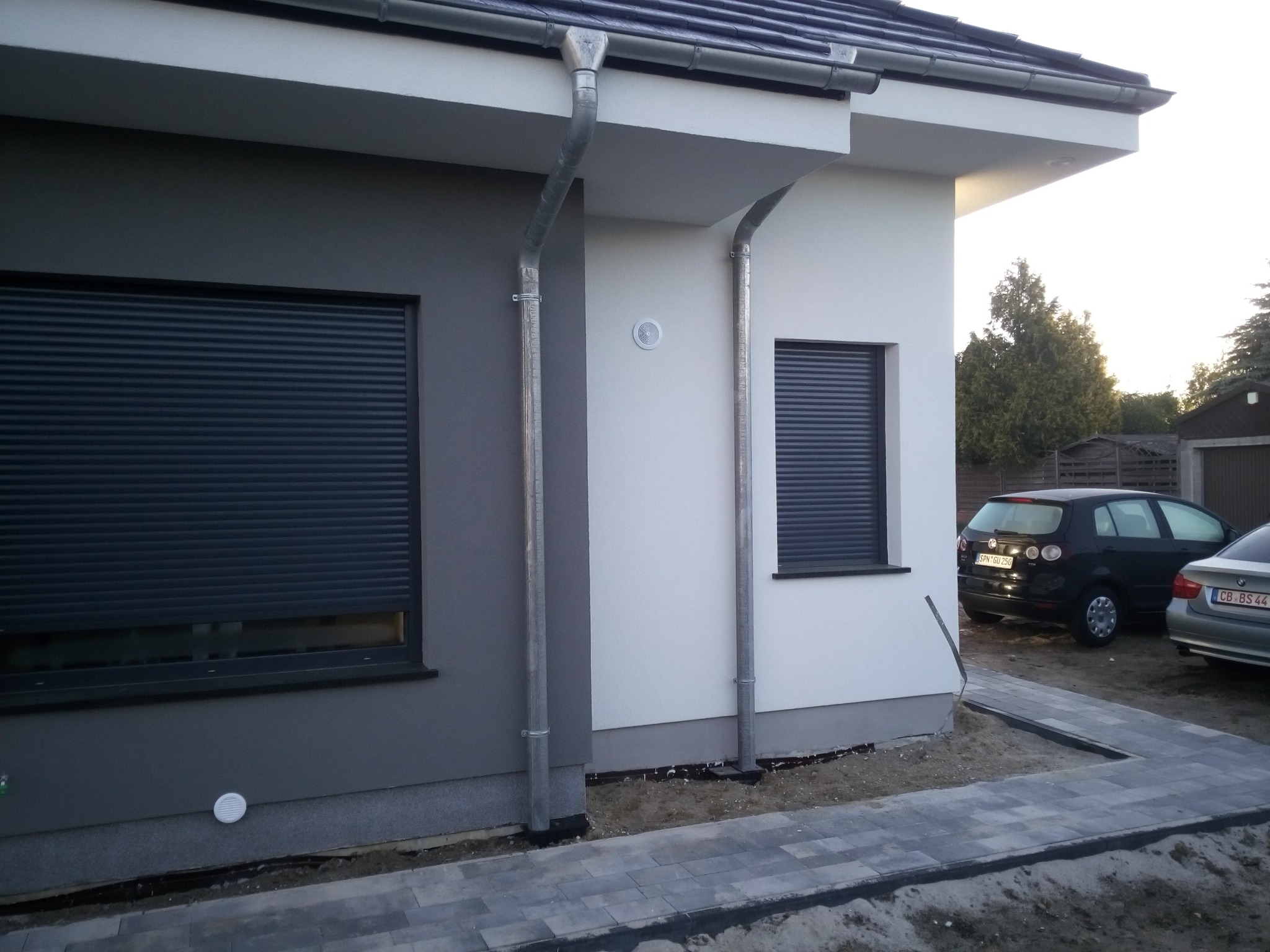 Dwukolorowa elewacja domu jednorodzinnego z szarymi roletami zewnętrznymi, widoczne rynny spustowe i częściowo ułożona kostka brukowa wokół budynku.