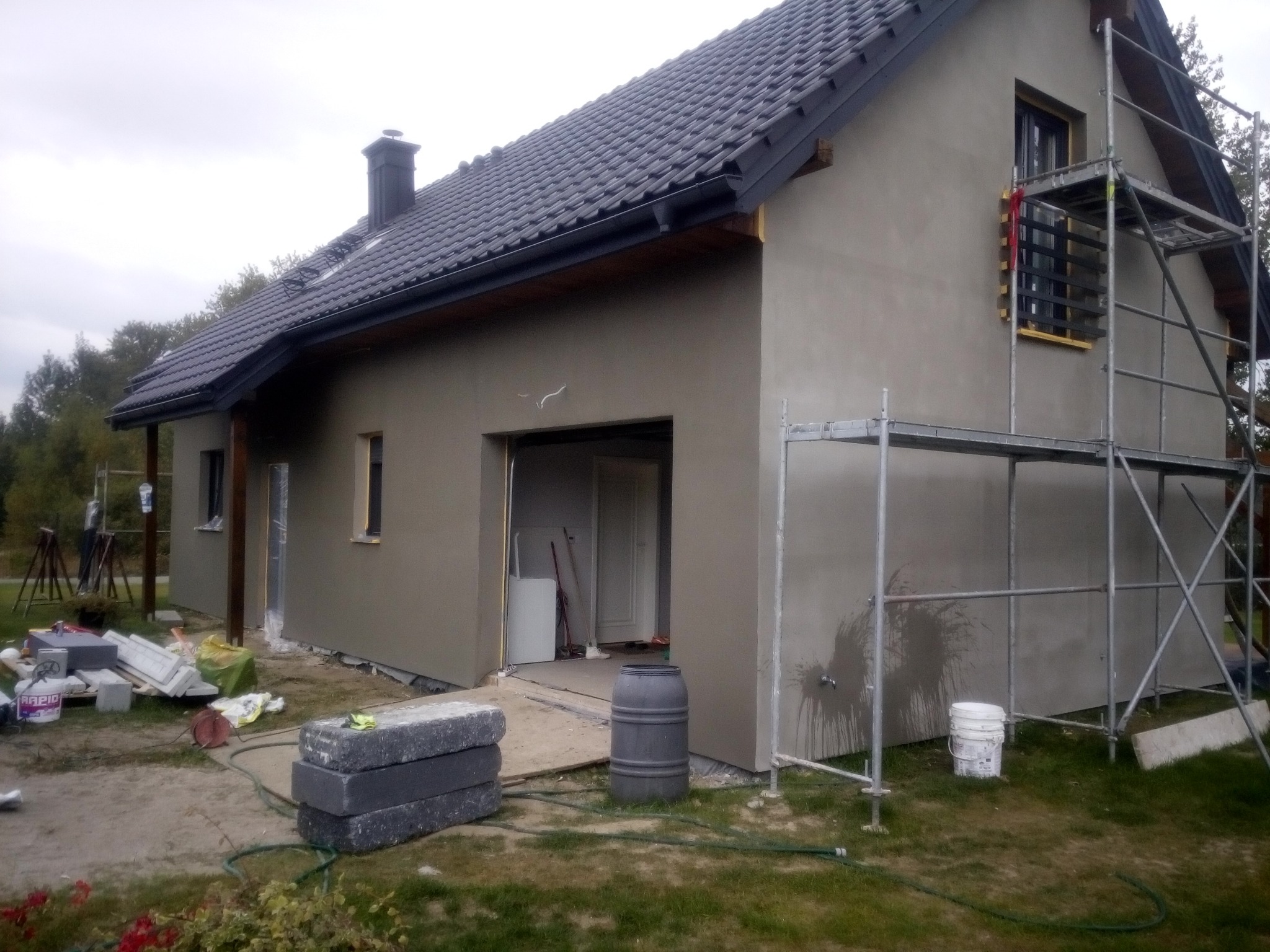 Dom jednorodzinny w trakcie prac elewacyjnych, widoczne rusztowanie, świeżo nałożona warstwa tynku oraz materiały budowlane na trawniku.