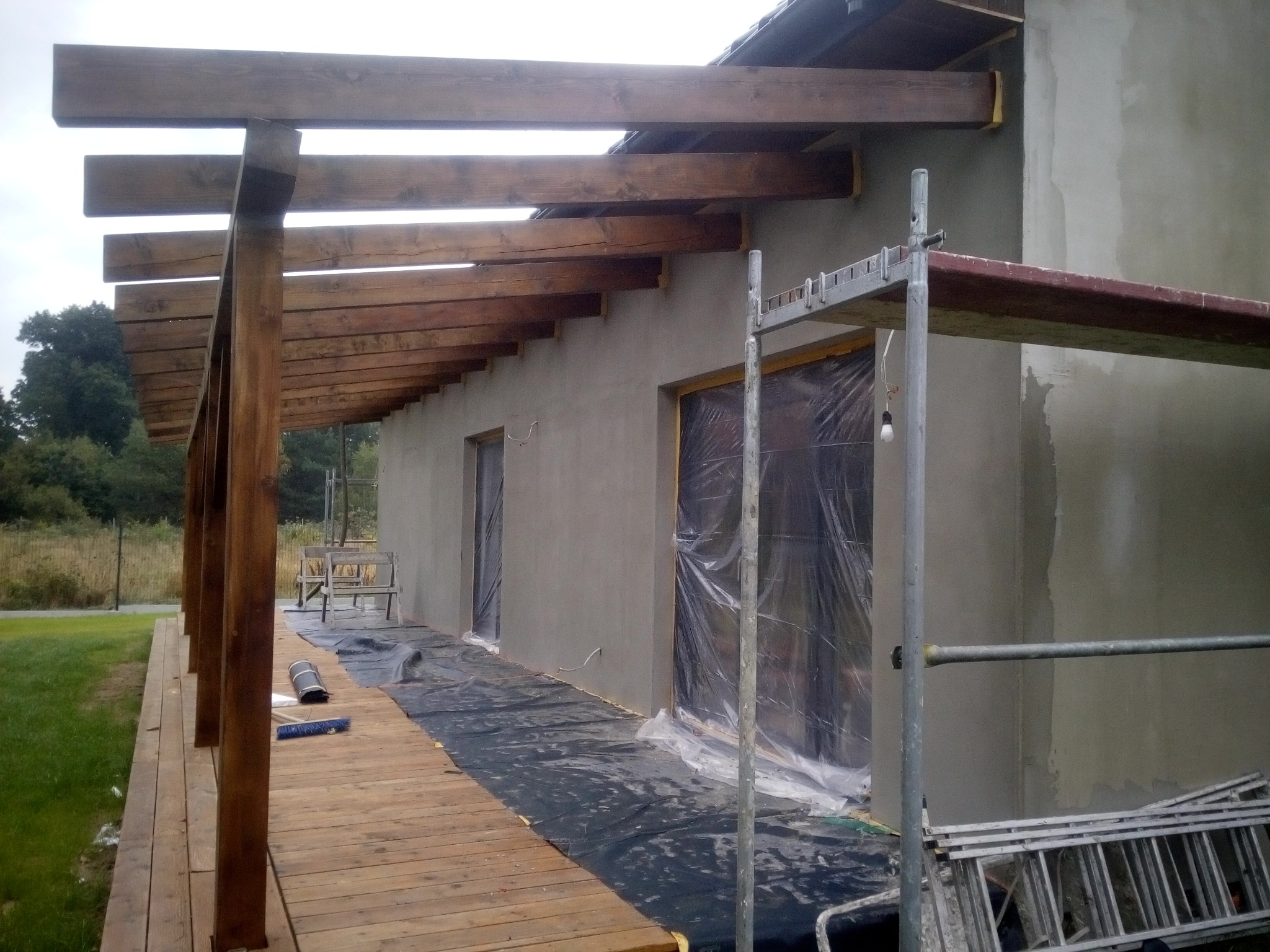 Surowa elewacja budynku z drewnianą pergolą w trakcie budowy, okna zabezpieczone folią, widoczne rusztowanie i narzędzia na drewnianym tarasie.