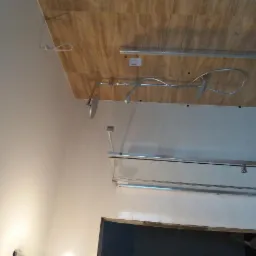 Zamontowana kabina prysznicowa ze szklanymi drzwiami i deszczownicą na suficie wyłożonym płytkami imitującymi drewno. Widoczne niedociągnięcia w wykończeniu.