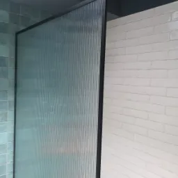 Kabina prysznicowa z ryflowanego szkła w czarnej ramie, przylegająca do ściany z białej cegły, oświetlona wiszącą żarówką.