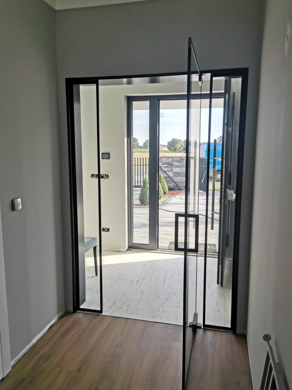 Nowoczesne szklane drzwi wewnętrzne z czarną ramą i podłużnymi uchwytami, widok na taras i ogród z elementami małej architektury.