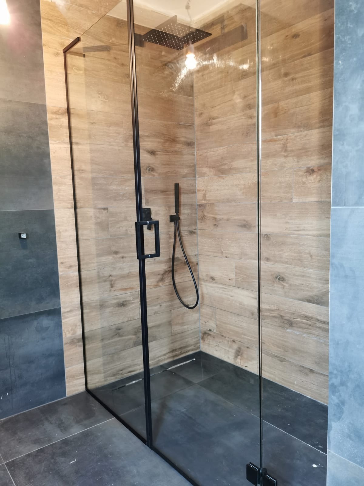 Nowoczesna kabina prysznicowa ze szklanymi ścianami i czarnymi profilami na tle płytek imitujących drewno i szarych płytek.