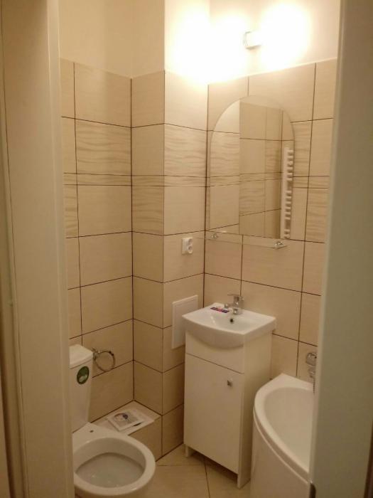 Wykończona łazienka w beżowych płytkach z poziomym wzorem, umywalka z szafką, toaleta z dokumentem na obudowie i owalne lustro nad umywalką.