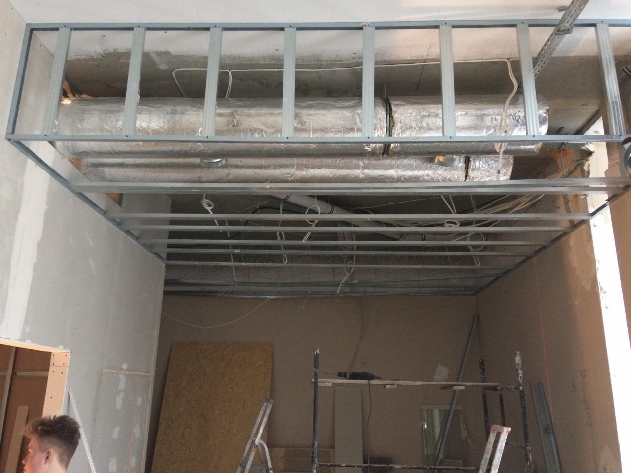 Metalowa konstrukcja sufitu podwieszanego z widocznymi przewodami i izolacją termiczną, w trakcie adaptacji lokalu, widoczny fragment ściany z płyt gipsowo-kartonowych.